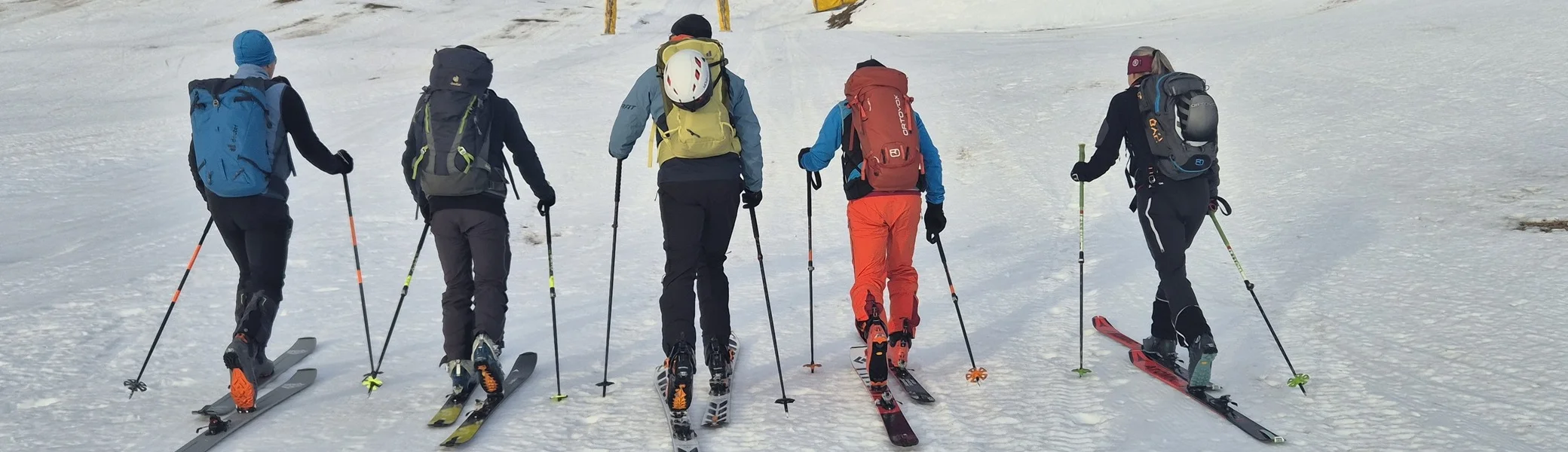 Fünf Skifahrer mit Rucksäcken steigen auf einer Schneepiste bergauf, im Hintergrund Hütten und Berge | © DAV-Fn / R. Lebedeff
