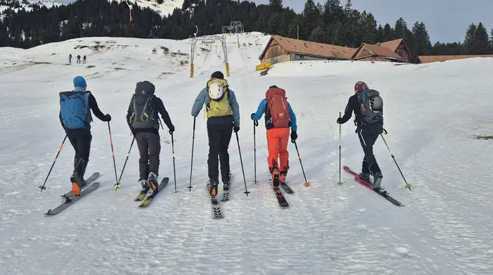 Fünf Skifahrer mit Rucksäcken steigen auf einer Schneepiste bergauf, im Hintergrund Hütten und Berge | © DAV-Fn / R. Lebedeff