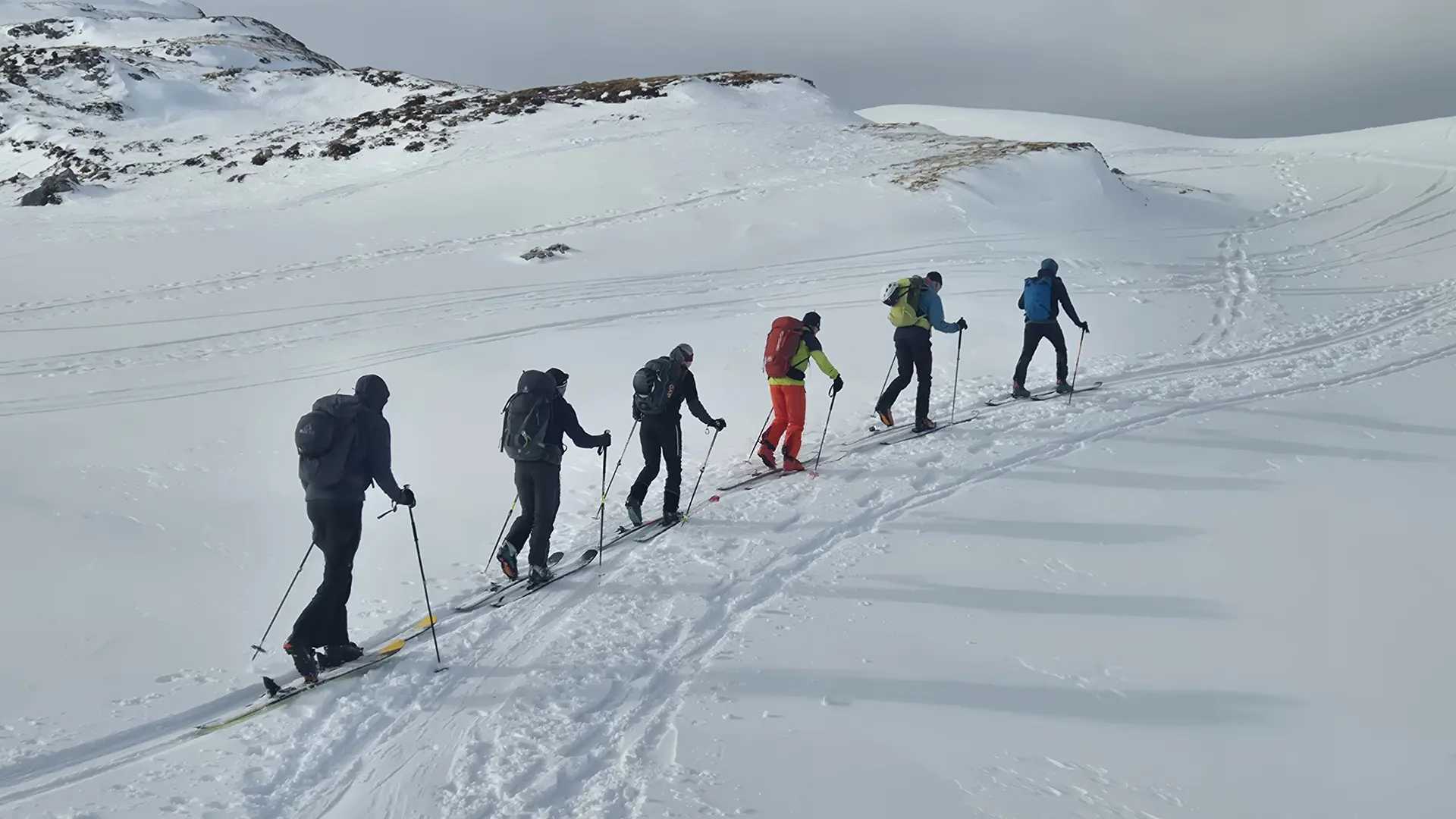 Sechs Skitourengeher in einer Reihe steigen auf verschneitem Berghang bei bewölktem Himmel auf | © DAV-Fn / R. Lebedeff