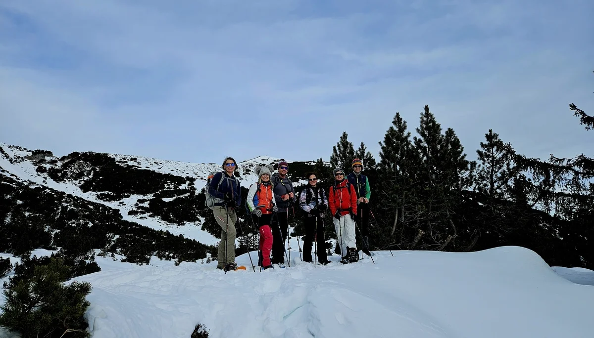 Gruppe von sechs Personen mit Wanderkleidung und Rucksäcken steht auf schneebedecktem Hang vor Nadelbäumen und Bergen | © DAV-FN / A. Schababerle
