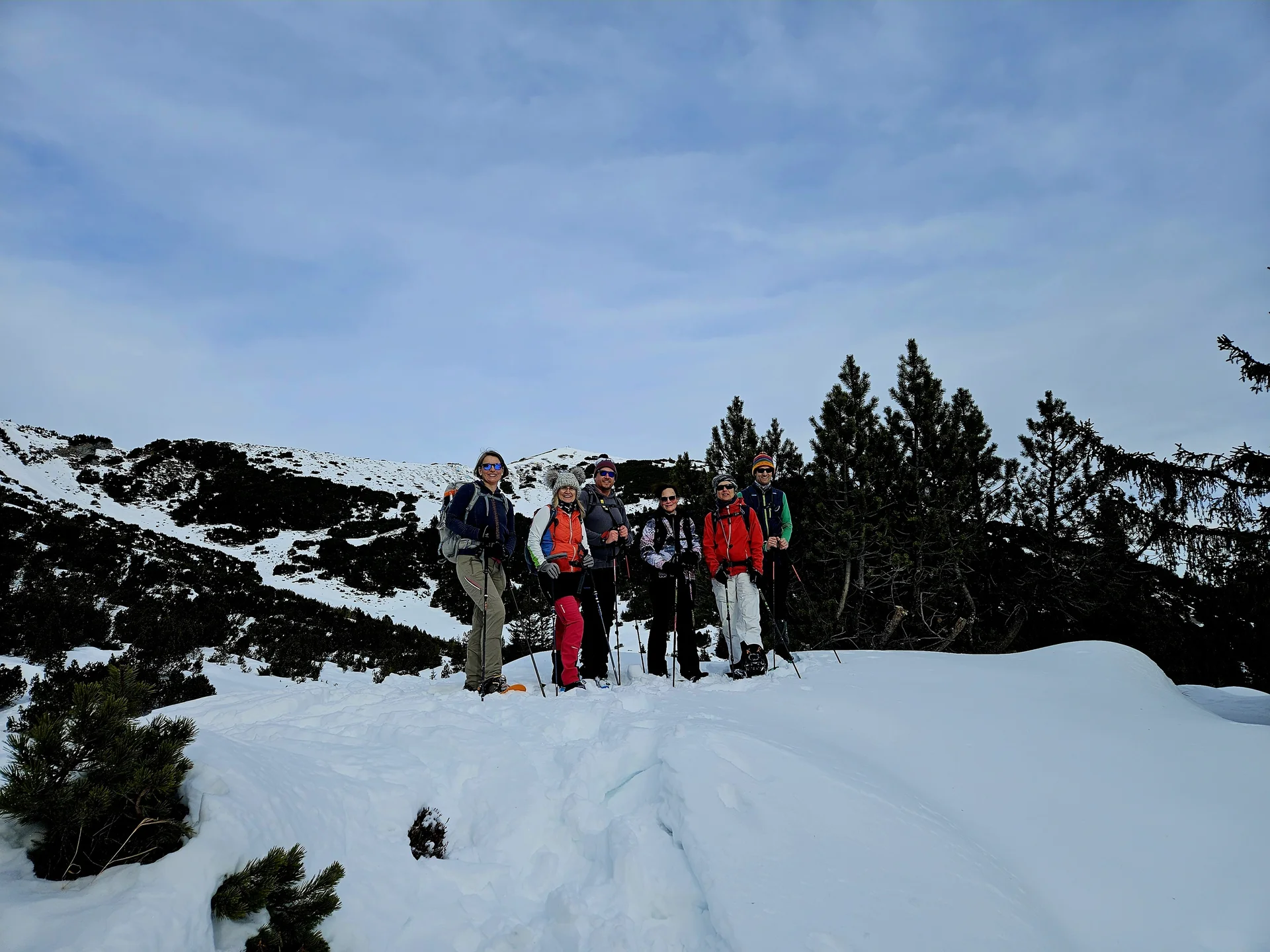 Gruppe von sechs Personen mit Wanderkleidung und Rucksäcken steht auf schneebedecktem Hang vor Nadelbäumen und Bergen | © DAV-FN / A. Schababerle