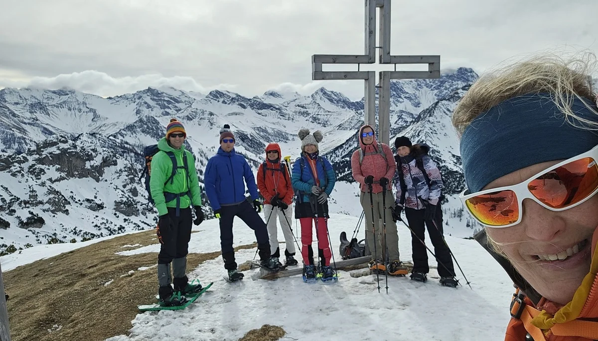 Gruppe von Wanderern mit Schneeschuhen steht auf einem Berggipfel neben einem großen Holzkreuz vor schneebedeckten Bergen | © DAV-FN / A. Schababerle