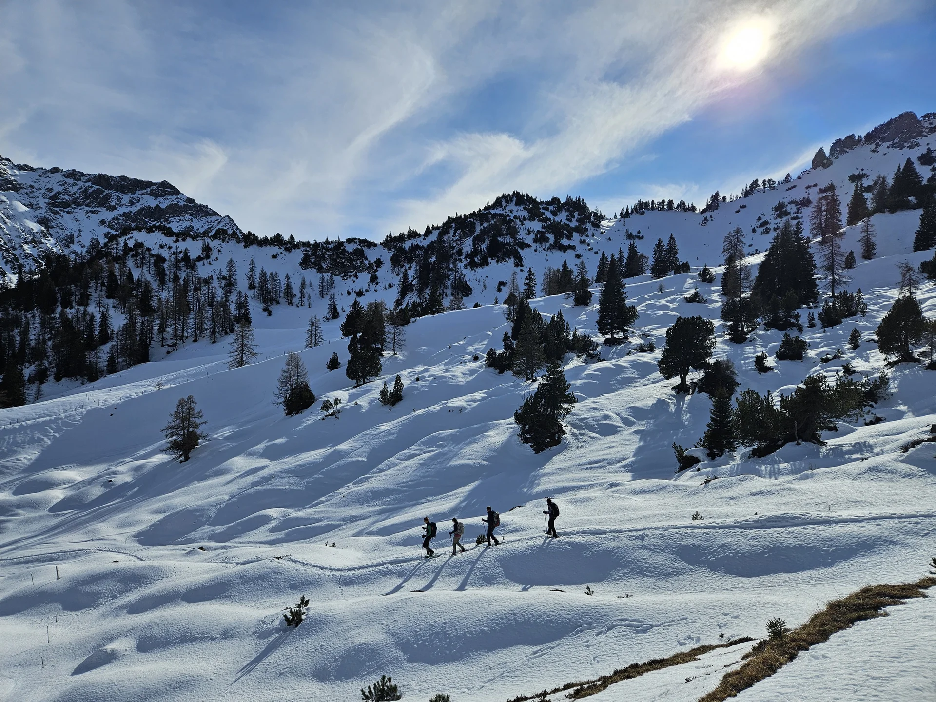 Vier Personen wandern in einer Reihe auf verschneitem Bergpfad unter bewölktem Himmel mit bewaldeten Berghängen im Hintergrund | © DAV-FN / A. Schababerle