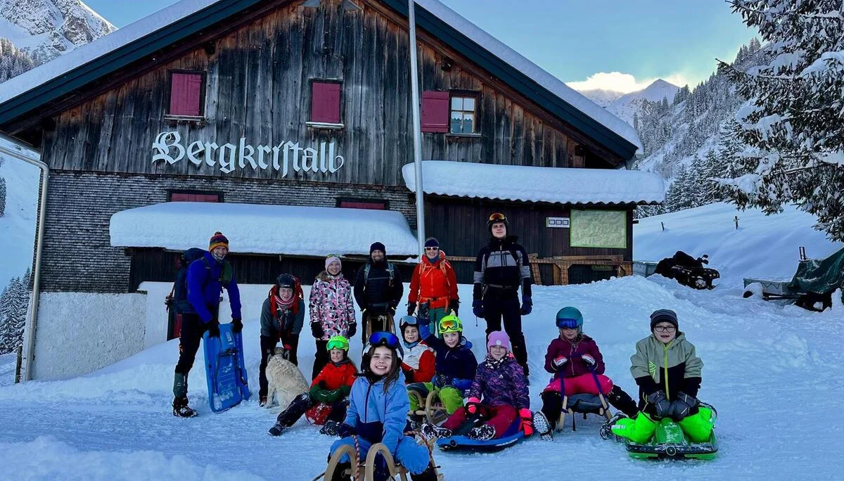 Gruppe von Kindern und Erwachsenen mit Schlitten vor einer Berghütte namens 'Bergkristall' in verschneiter Berglandschaft | © DAV-FN / A. Schababerle