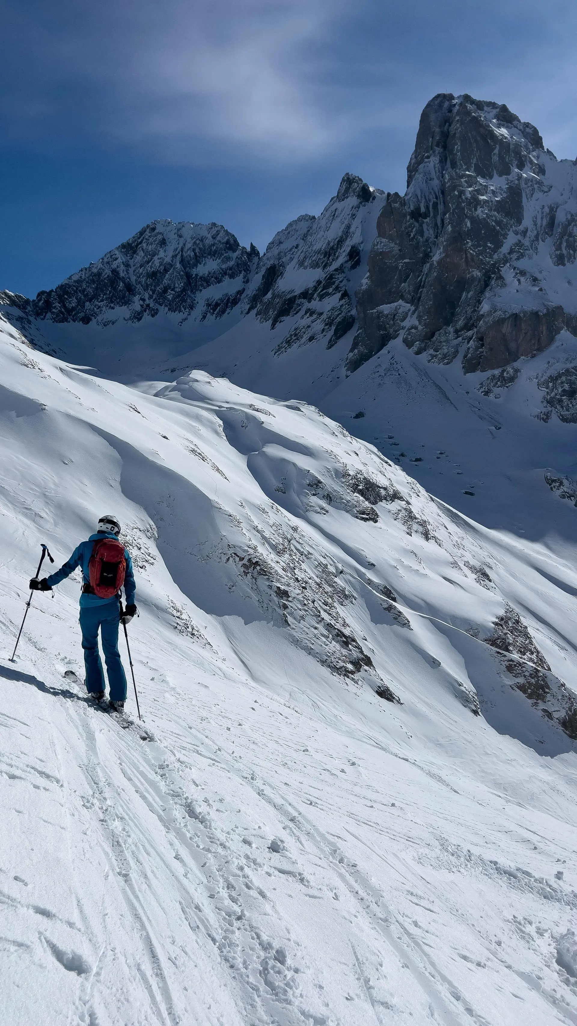 Skifahrer mit rotem Rucksack und blauer Kleidung auf verschneitem Hang vor steilen, schroffen Berggipfeln | © DAV-FN / S. Schmidhuber
