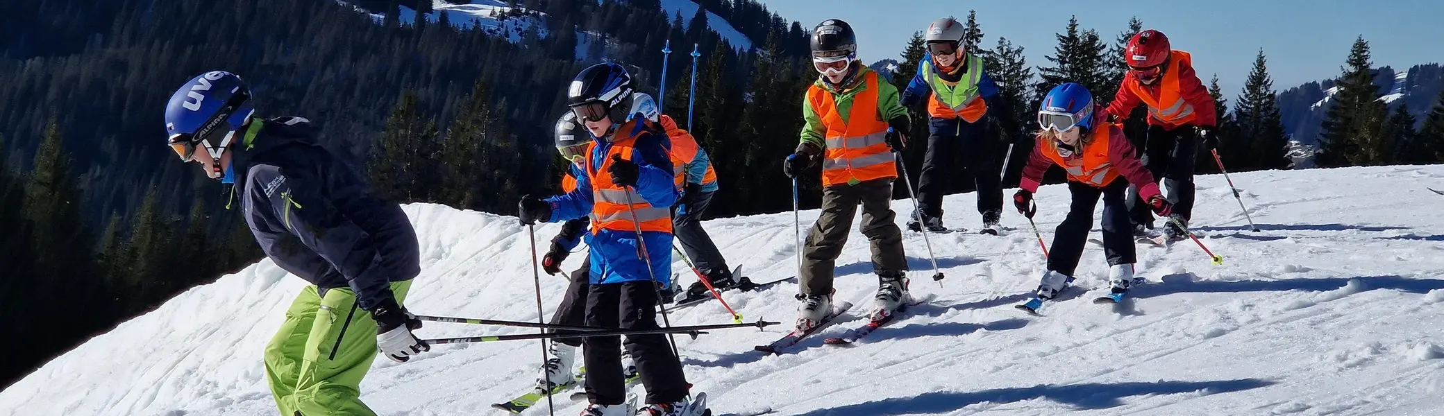 Gruppe von Kindern mit Skihelmen und Skistöcken beim Skifahren auf einer schneebedeckten Piste vor bewaldeten Bergen | © DAV-FN / J. Kastens