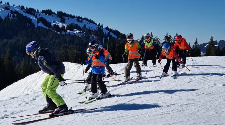 Gruppe von Kindern mit Skihelmen und Skistöcken beim Skifahren auf einer schneebedeckten Piste vor bewaldeten Bergen | © DAV-FN / J. Kastens