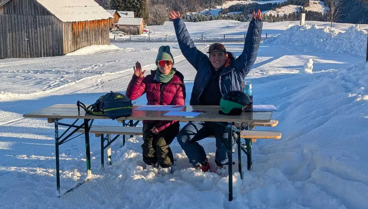 Zwei Personen sitzen an einem Holztisch im Schnee vor einer verschneiten Hütte und heben die Arme | © DAV-FN / L. Eisele