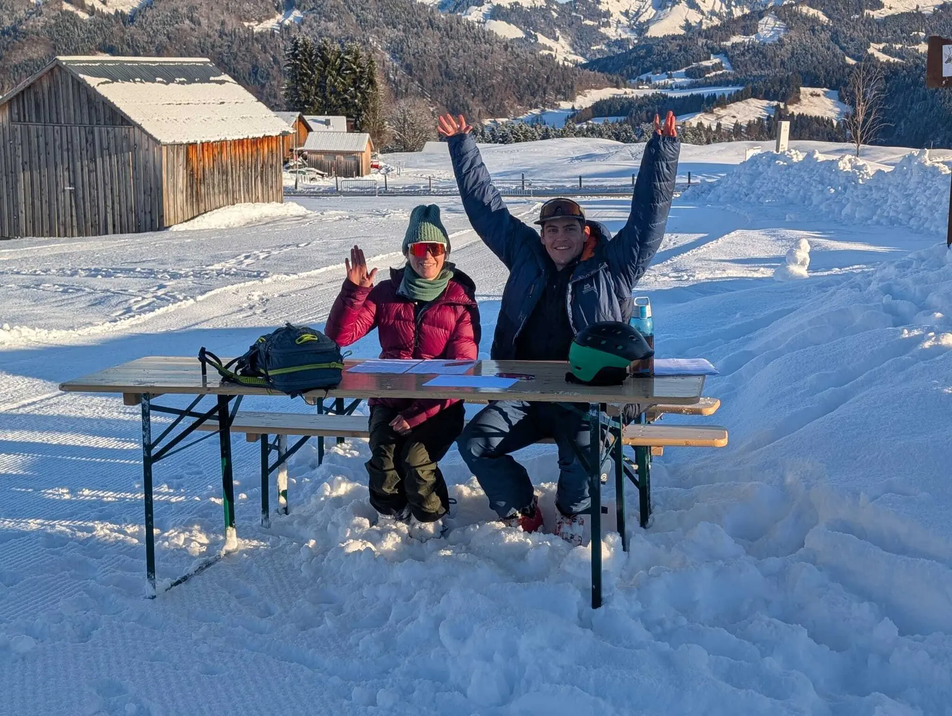Zwei Personen sitzen an einem Holztisch im Schnee vor einer verschneiten Hütte und heben die Arme | © DAV-FN / L. Eisele