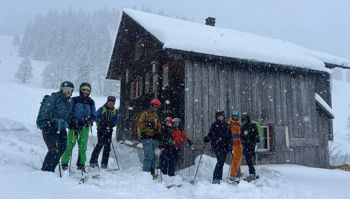 Gruppe von sieben Personen mit Skiausrüstung steht vor einer verschneiten Holzhütte in winterlicher Berglandschaft | © DAV-FN / C. + C. Ritter