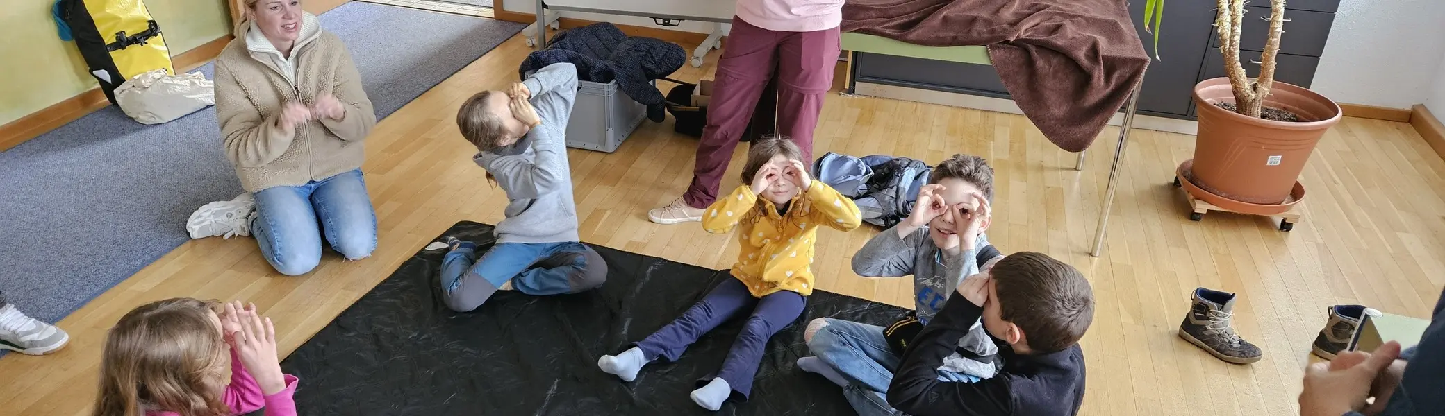 Kinder sitzen im Kreis auf dem Boden eines Raums und eine stehende Person spricht zu ihnen | © DAV-FN / A. Schababerle