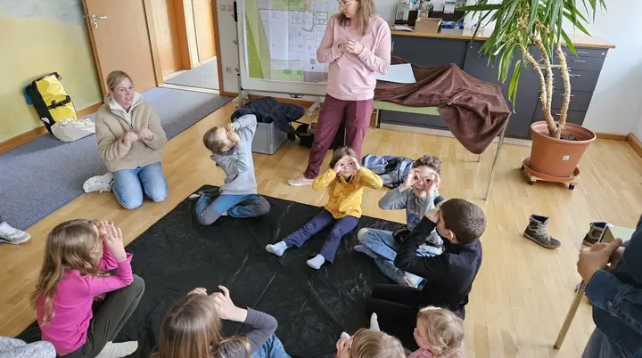 Kinder sitzen im Kreis auf dem Boden eines Raums und eine stehende Person spricht zu ihnen | © DAV-FN / A. Schababerle