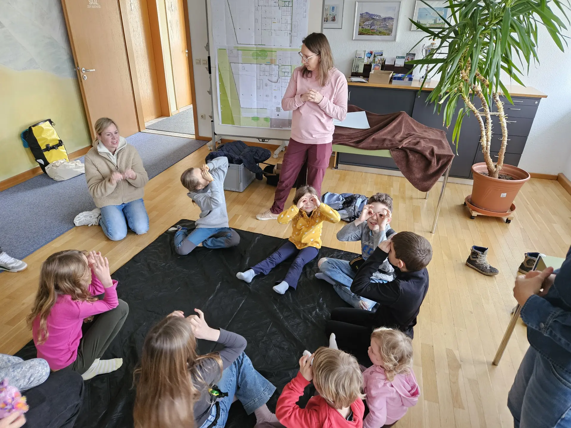Kinder sitzen im Kreis auf dem Boden eines Raums und eine stehende Person spricht zu ihnen | © DAV-FN / A. Schababerle