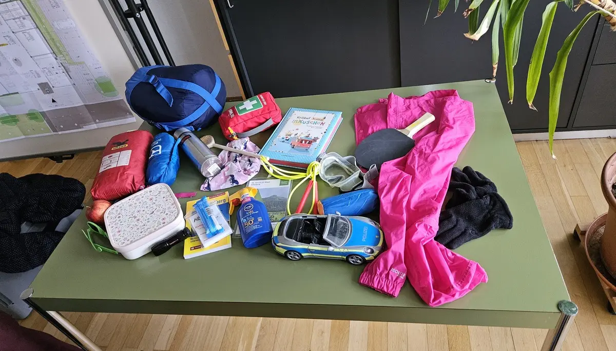 Auf einem grünen Tisch liegen verschiedene Gegenstände wie pinke Hose, Spielzeugauto, Erste-Hilfe-Set, Bücher und weitere persönliche Utensilien | © DAV-FN / A. Schababerle