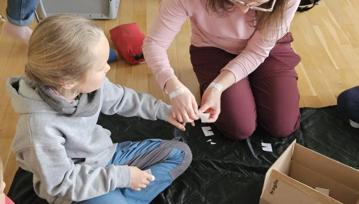 Eine Frau mit Brille und rosa Pullover sitzt auf dem Boden und hält die Hand eines Kindes, das auf einem schwarzen Tuch sitzt, neben einer offenen Pappschachtel. | © DAV-FN / A. Schababerle