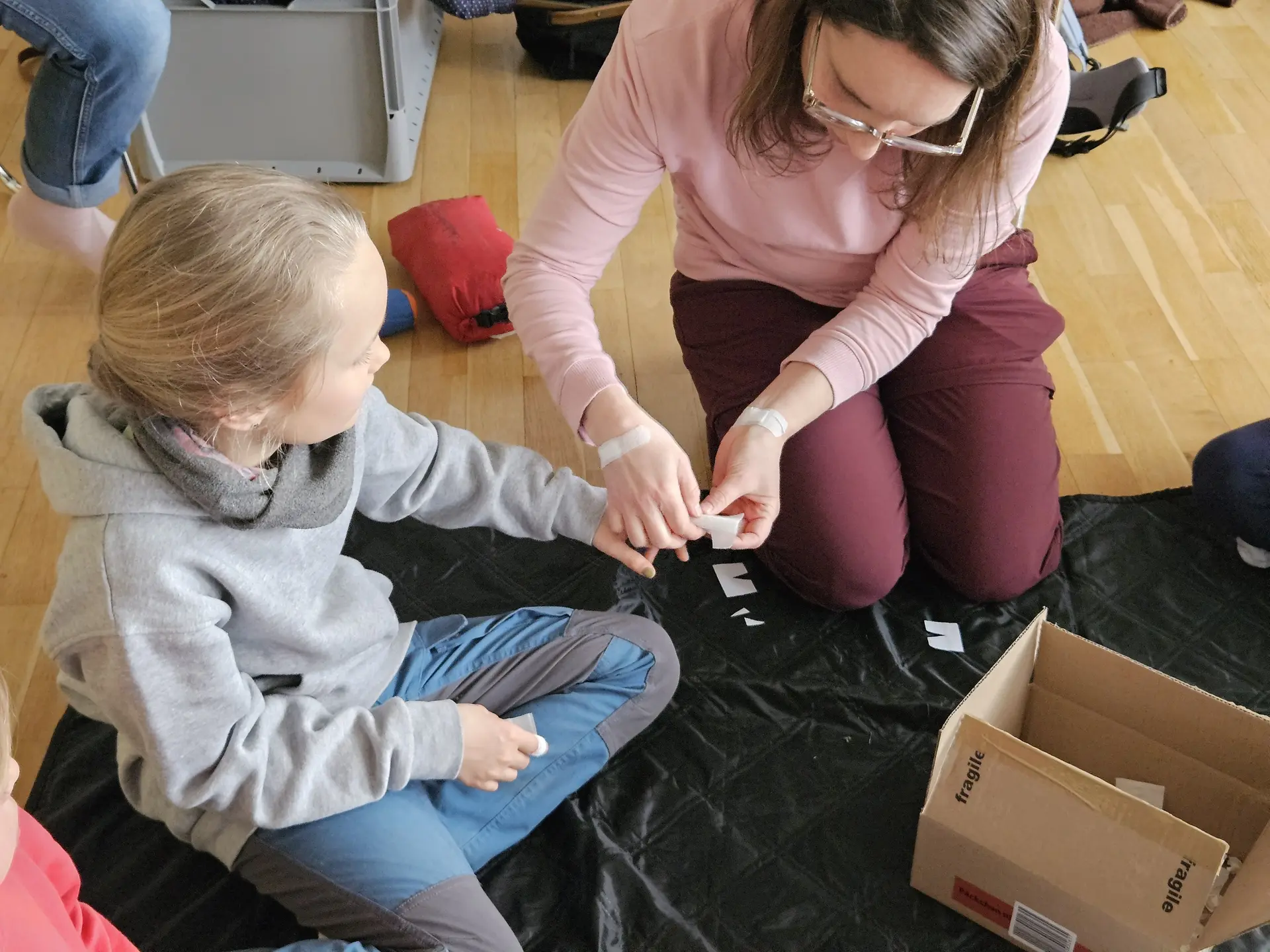 Eine Frau mit Brille und rosa Pullover sitzt auf dem Boden und hält die Hand eines Kindes, das auf einem schwarzen Tuch sitzt, neben einer offenen Pappschachtel. | © DAV-FN / A. Schababerle