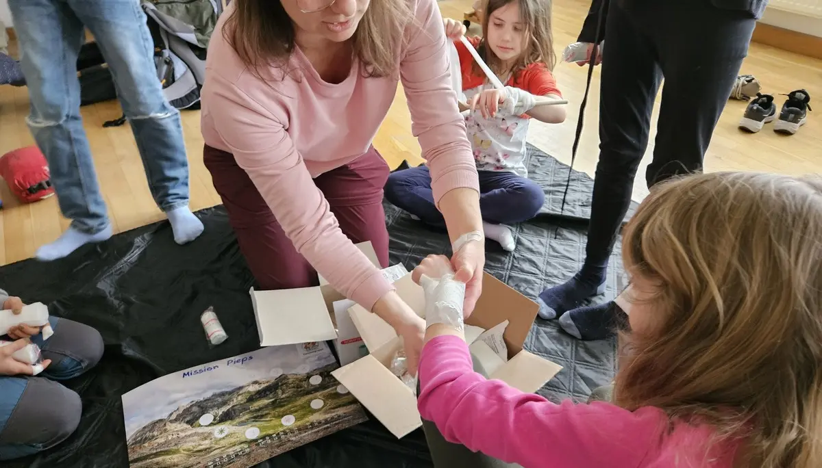 Mehrere Personen sitzen auf dem Boden, eine Person verbindet den Arm eines Kindes mit einer Bandage | © DAV-FN / A. Schababerle