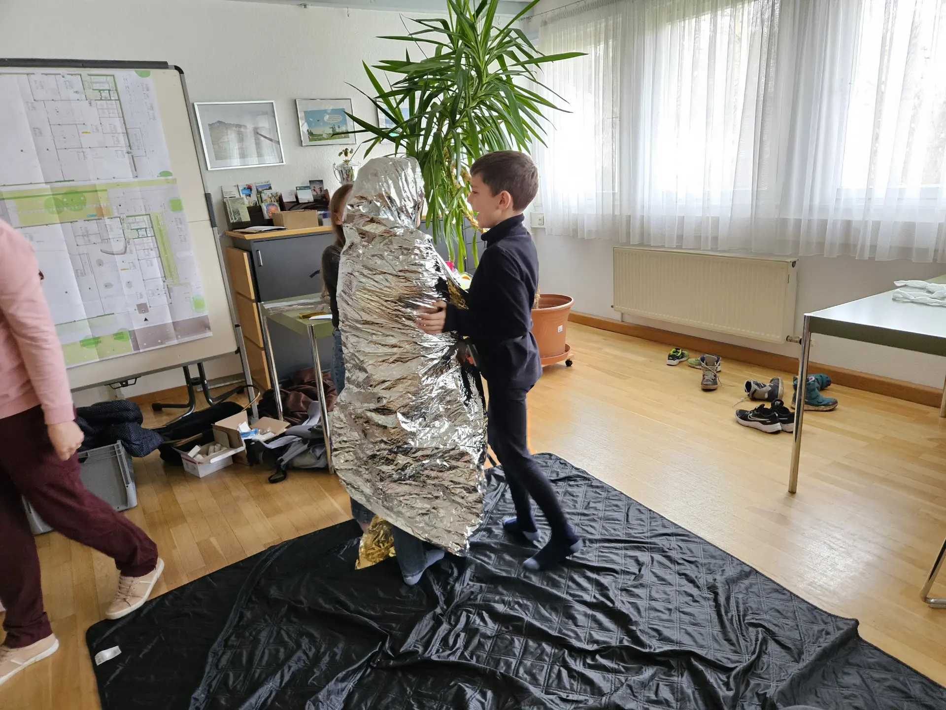 Kind steht vor einer Person, die in eine silberne Rettungsdecke eingewickelt ist, in einem hellen Raum mit Pflanzen und Möbeln | © DAV-FN / A. Schababerle