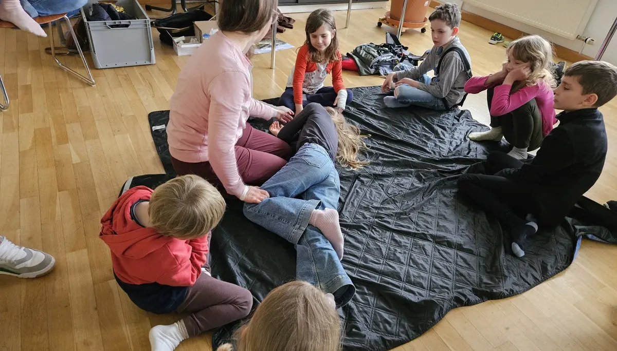 Gruppe von Kindern sitzt auf dem Boden um eine Frau, die ein Kind in die Stabile Seitenlage dreht, in einem hellen Raum mit Holzboden | © DAV-FN / A. Schababerle