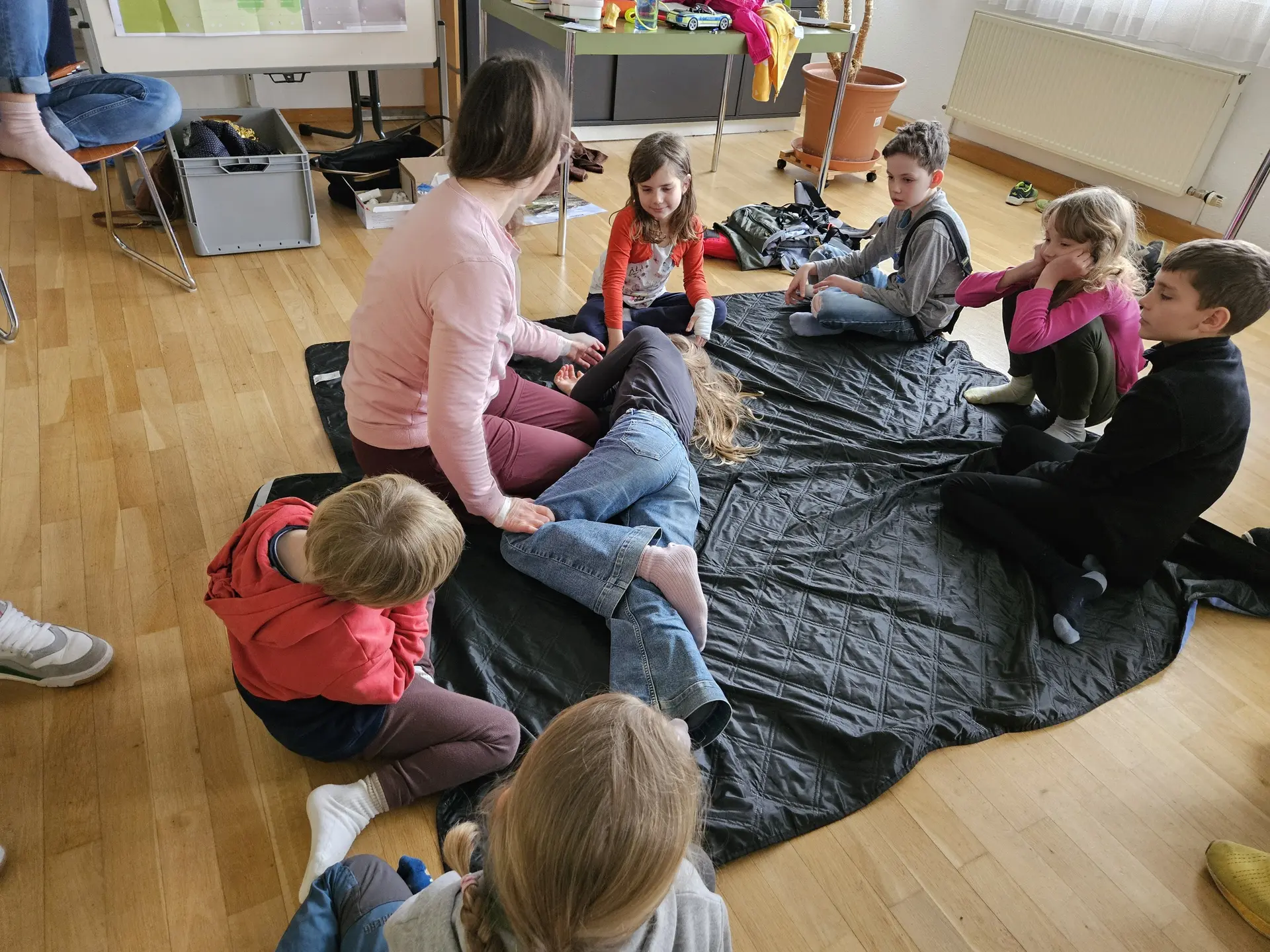 Gruppe von Kindern sitzt auf dem Boden um eine Frau, die ein Kind in die Stabile Seitenlage dreht, in einem hellen Raum mit Holzboden | © DAV-FN / A. Schababerle