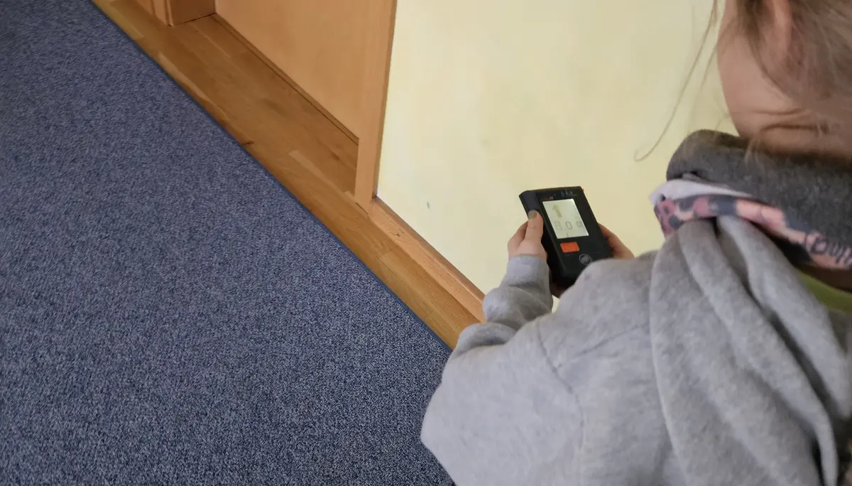 Kind von hinten mit grauem Kapuzenpullover hält ein Lawinenverschüttetensuchgerät in der Hand und steht auf blauem Teppich vor einer hellen Wand | © DAV-FN / A. Schababerle