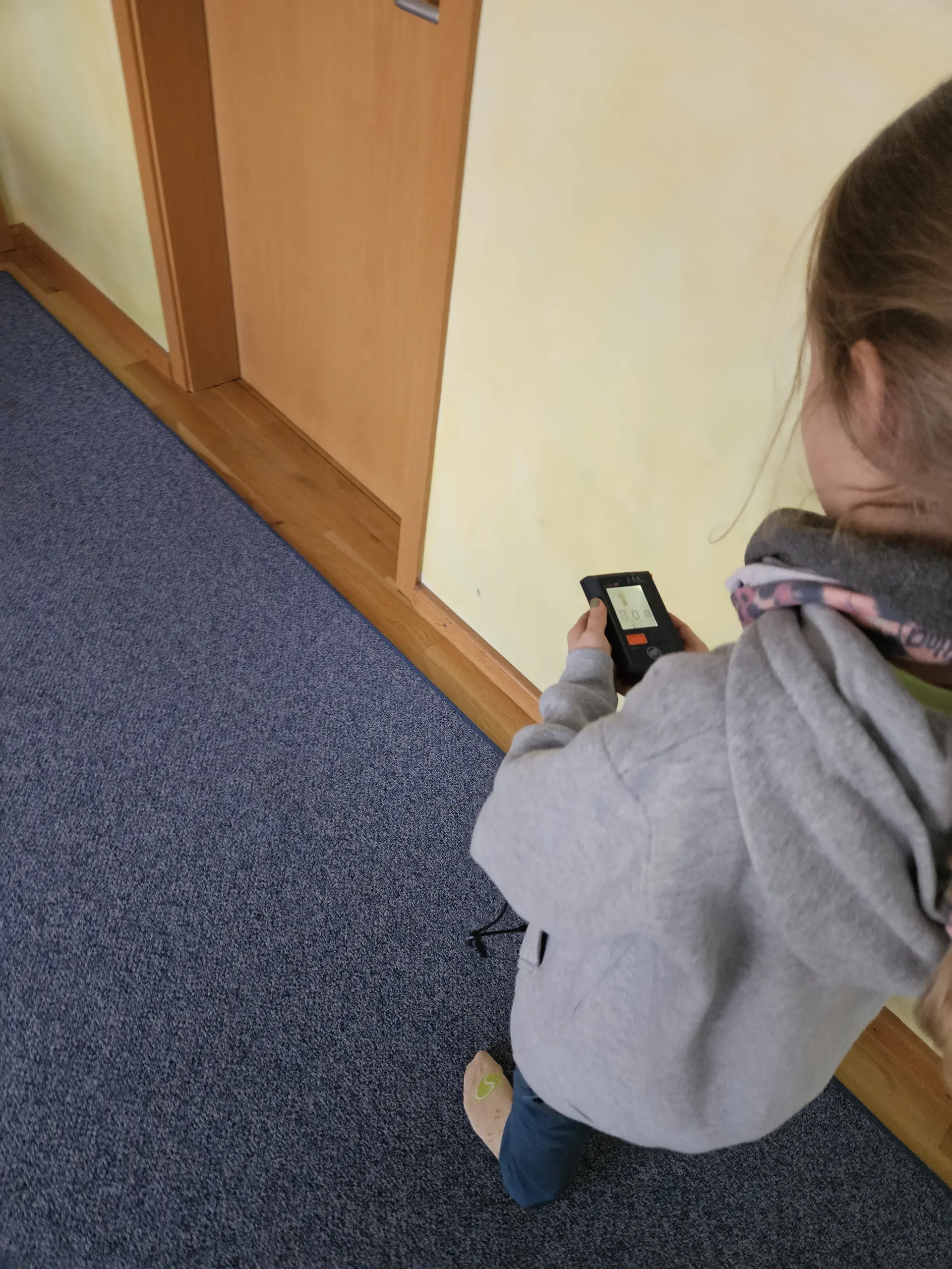 Kind von hinten mit grauem Kapuzenpullover hält ein Lawinenverschüttetensuchgerät in der Hand und steht auf blauem Teppich vor einer hellen Wand | © DAV-FN / A. Schababerle