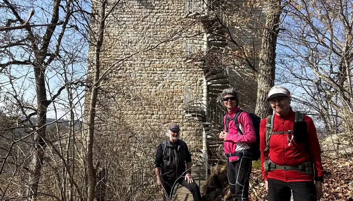 Drei Wanderer mit Rucksäcken stehen auf einem felsigen Waldweg vor einem hohen, rechteckigen Steinturm mit außenliegender Wendeltreppe | © DAV-FN / A. Schech