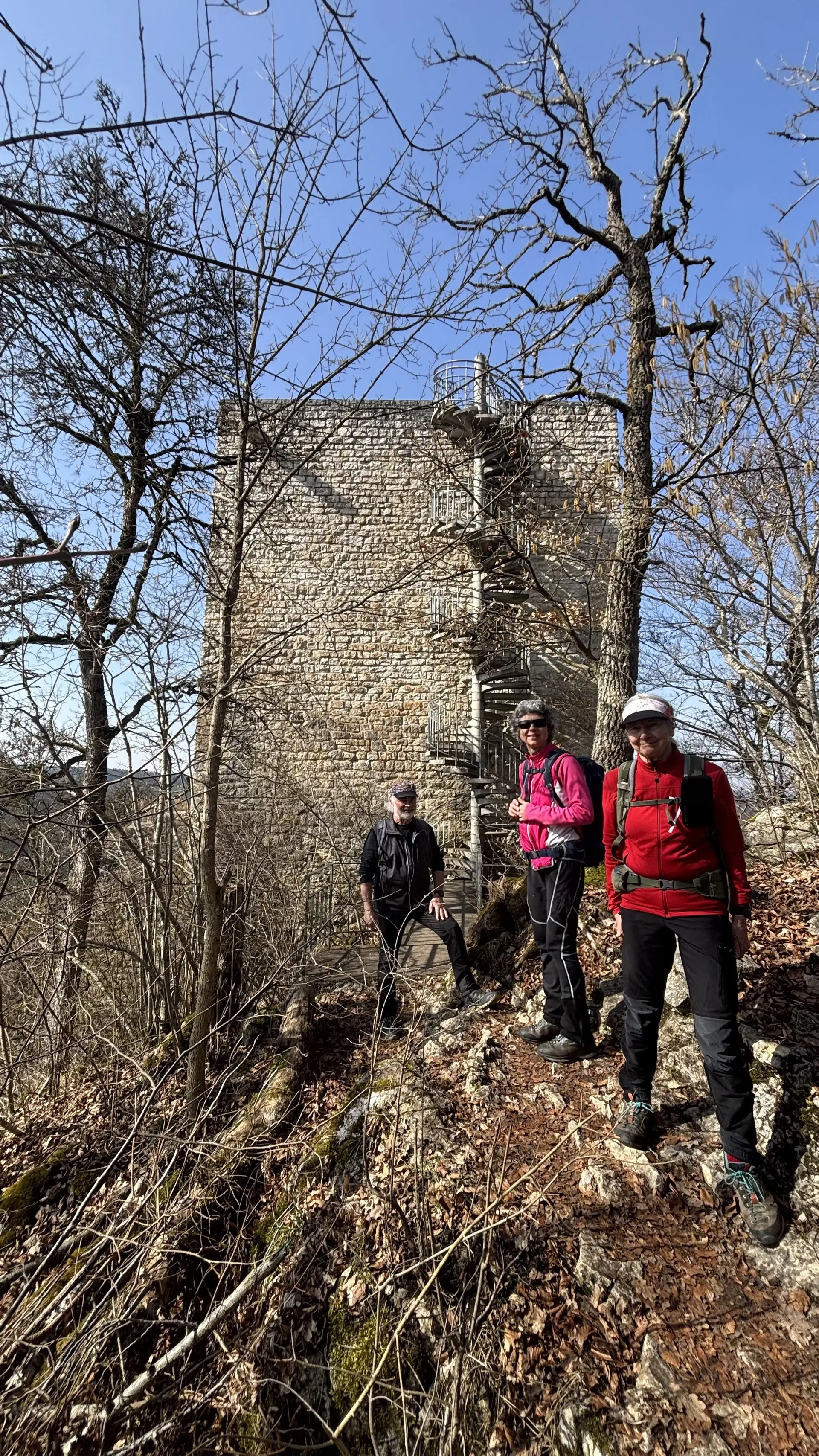 Drei Wanderer mit Rucksäcken stehen auf einem felsigen Waldweg vor einem hohen, rechteckigen Steinturm mit außenliegender Wendeltreppe | © DAV-FN / A. Schech