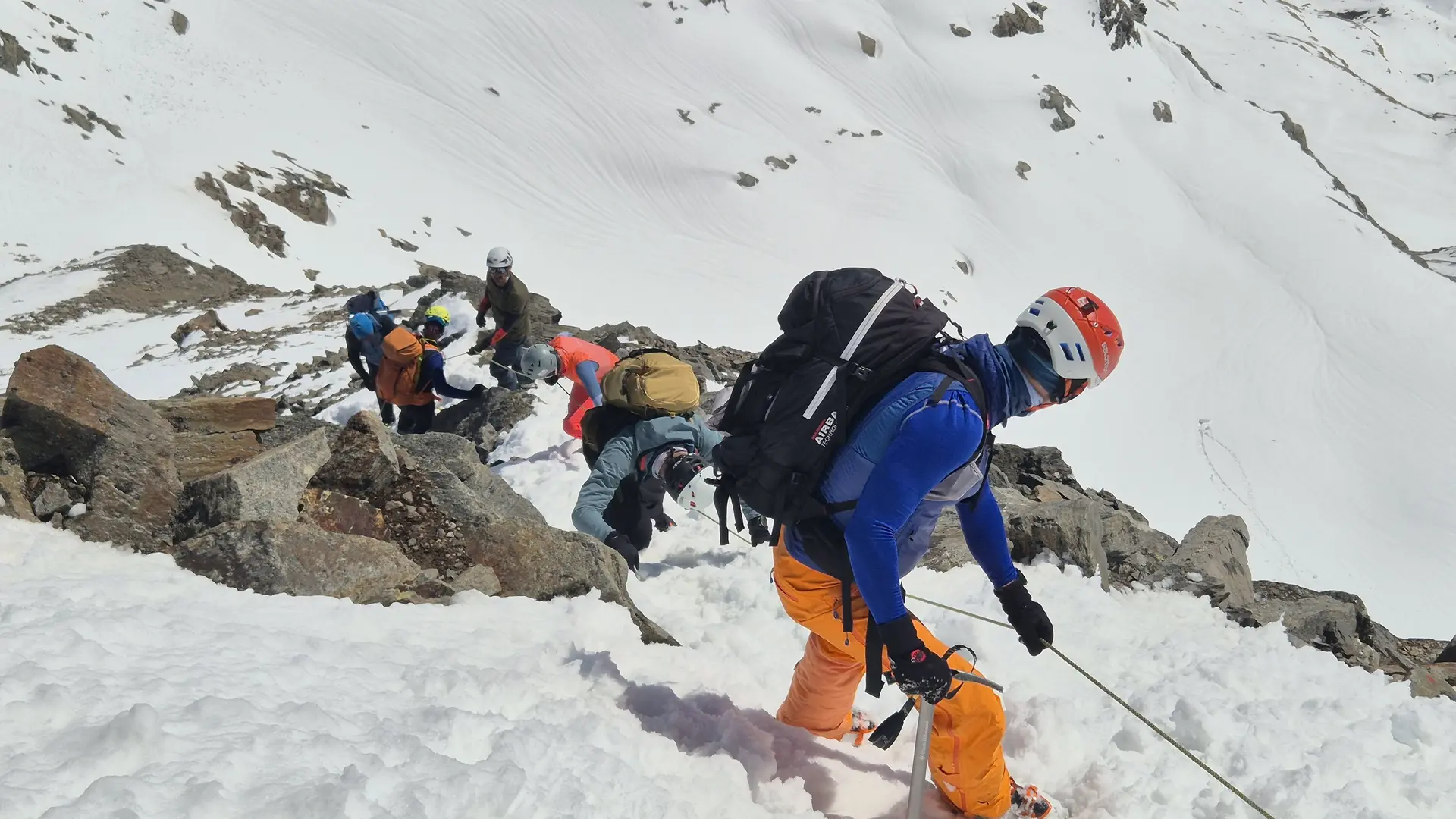 Gruppe von Bergsteigern mit Helmen und Rucksäcken klettert gesichert mit einem Fixseil einem schneebedeckten und felsendurchsetzten Berghang hinab. | © DAV-FN / R. Lebedeff