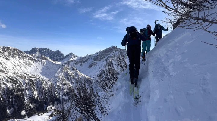 Drei Skitourengeher steigen auf einem schmalen, schneebedeckten Bergpfad mit Bergpanorama im Hintergrund auf | © DAV/Sandra Meyer