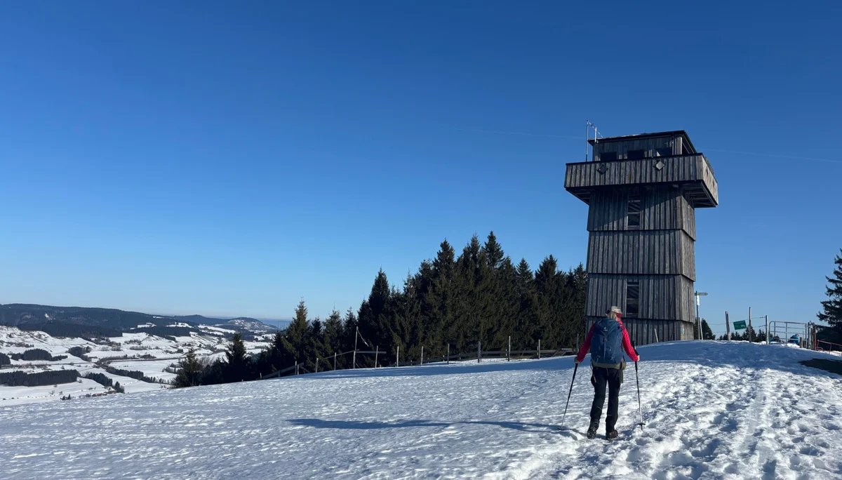 Person mit Wanderstöcken geht auf verschneitem Weg zu einem hölzernen Aussichtsturm auf einem Hügel mit Nadelbäumen im Hintergrund | © DAV-FN / A. Schech