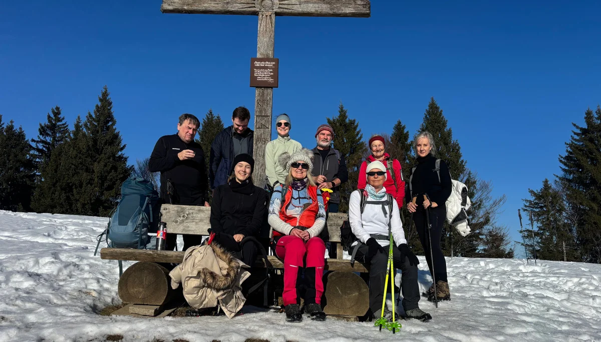 Gruppe von Wanderern mit Rucksäcken und Wanderstöcken sitzt und steht vor einem großen Holzkreuz auf schneebedecktem Boden mit Nadelbäumen im Hintergrund | © DAV-FN / A. Schech