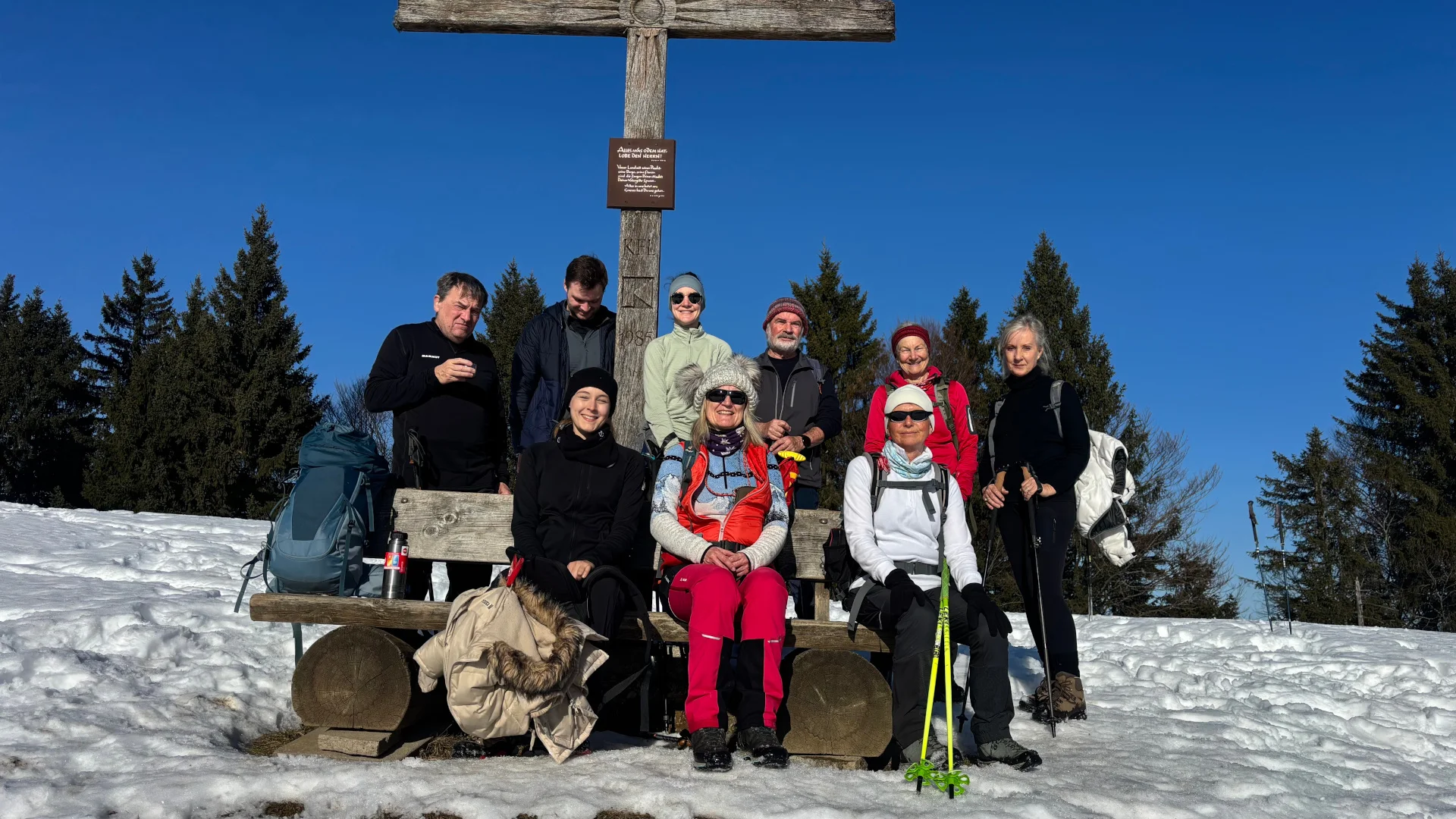 Gruppe von Wanderern mit Rucksäcken und Wanderstöcken sitzt und steht vor einem großen Holzkreuz auf schneebedecktem Boden mit Nadelbäumen im Hintergrund | © DAV-FN / A. Schech