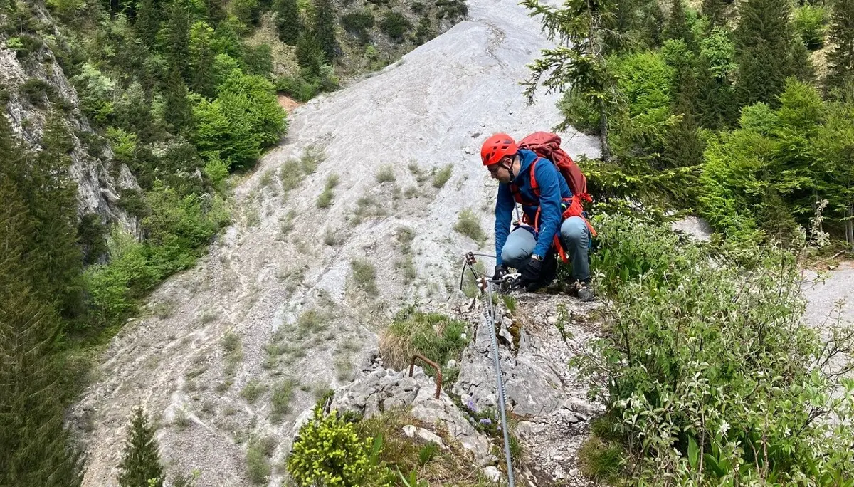 Bergsteiger mit Helm und Rucksack klettert an einem steilen Felsabhang in einem bewaldeten Gebirgstal | © DAV-FN / L. Fischer