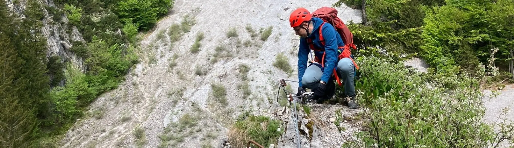 Bergsteiger mit Helm und Rucksack klettert an einem steilen Felsabhang in einem bewaldeten Gebirgstal | © DAV-FN / L. Fischer