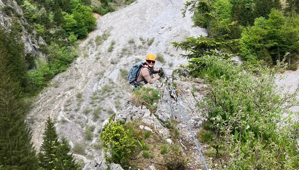 Bergsteiger mit Helm und Rucksack klettert an einem Stahlseil entlang eines steilen Felsgrats in einem bewaldeten Gebirgstal | © DAV-FN / L. Fischer