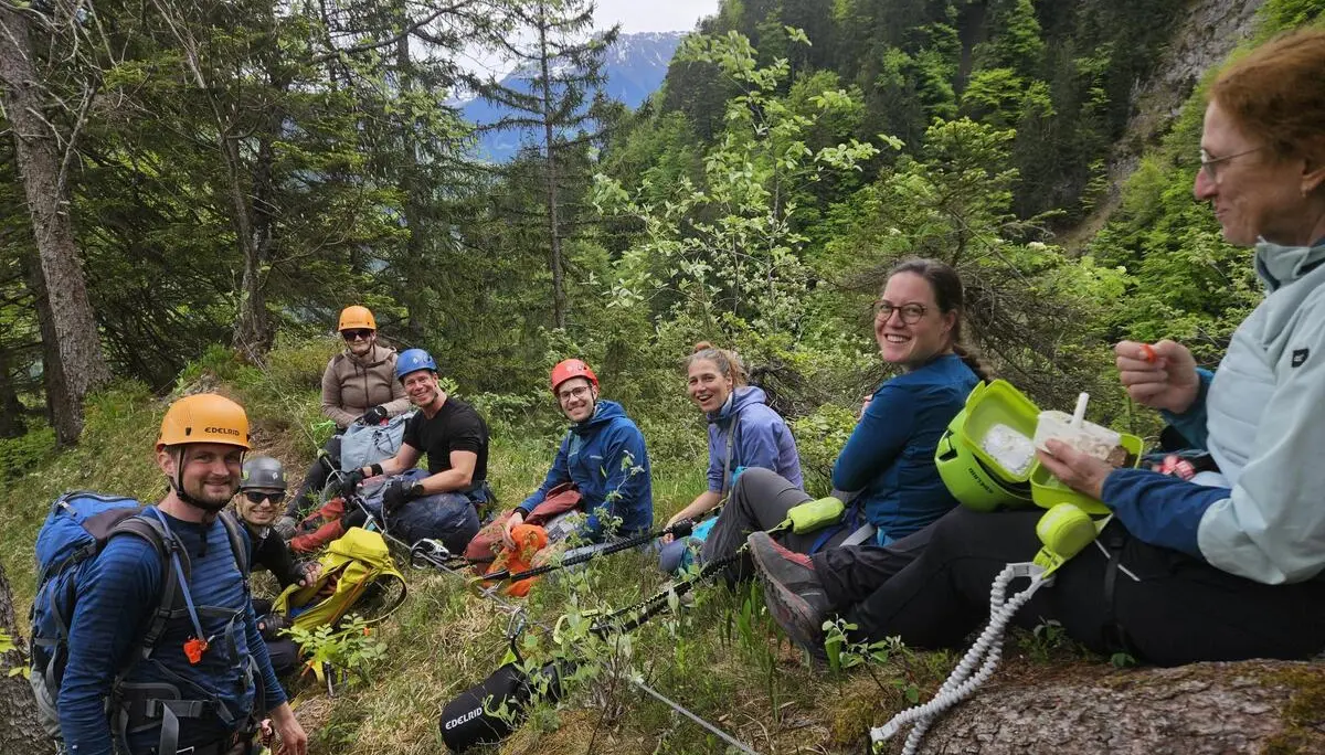 Gruppe von Kletterern mit Helmen und Rucksäcken sitzt auf einer Wiese und einem Baumstamm in einem bewaldeten Bergtal | © DAV-FN / S. Meyer