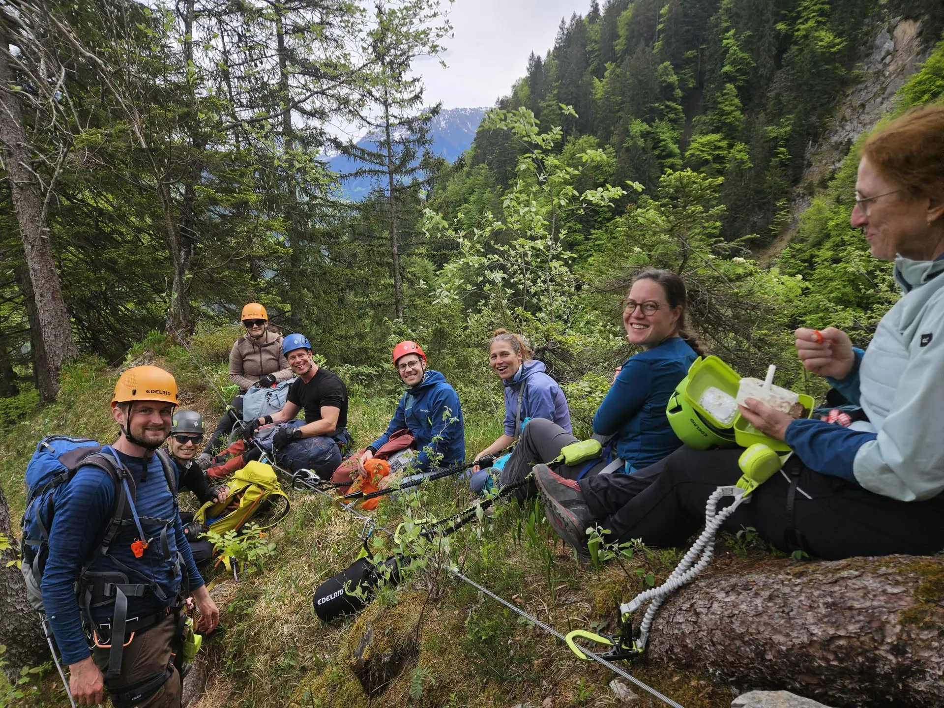 Gruppe von Kletterern mit Helmen und Rucksäcken sitzt auf einer Wiese und einem Baumstamm in einem bewaldeten Bergtal | © DAV-FN / S. Meyer