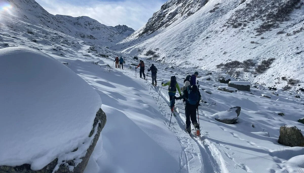 Gruppe von Skitourengehern in verschneitem Bergtal unter blauem Himmel mit Sonne und Wolken. | © DAV-FN / B. Rayk
