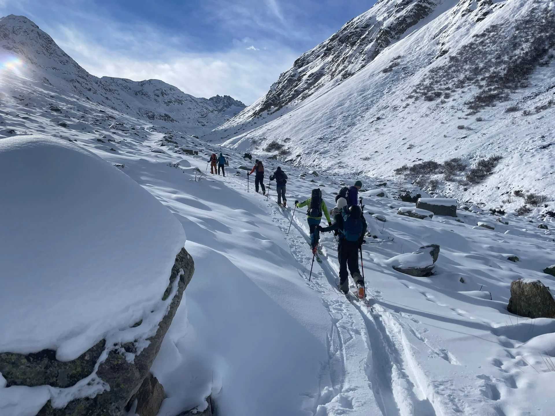 Gruppe von Skitourengehern in verschneitem Bergtal unter blauem Himmel mit Sonne und Wolken. | © DAV-FN / B. Rayk