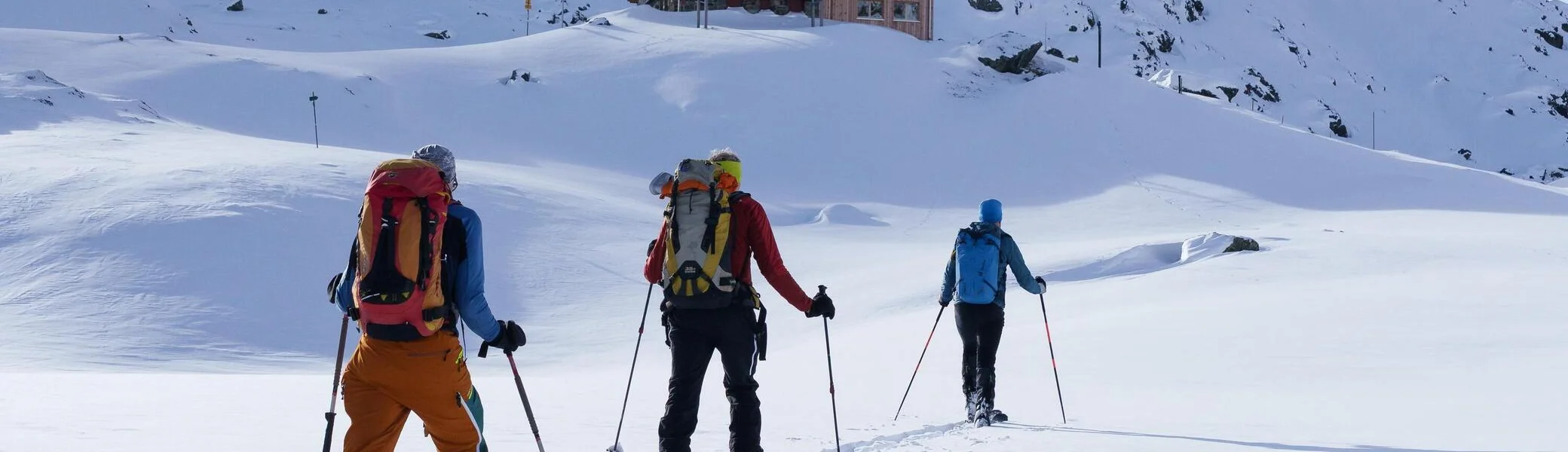 Drei Skitourengeher mit Rucksäcken und Skistöcken wandern in einer verschneiten Berglandschaft auf eine Berghütte zu. | © DAV-FN / B. Rayk