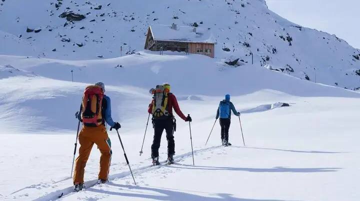 Drei Skitourengeher mit Rucksäcken und Skistöcken wandern in einer verschneiten Berglandschaft auf eine Berghütte zu. | © DAV-FN / B. Rayk
