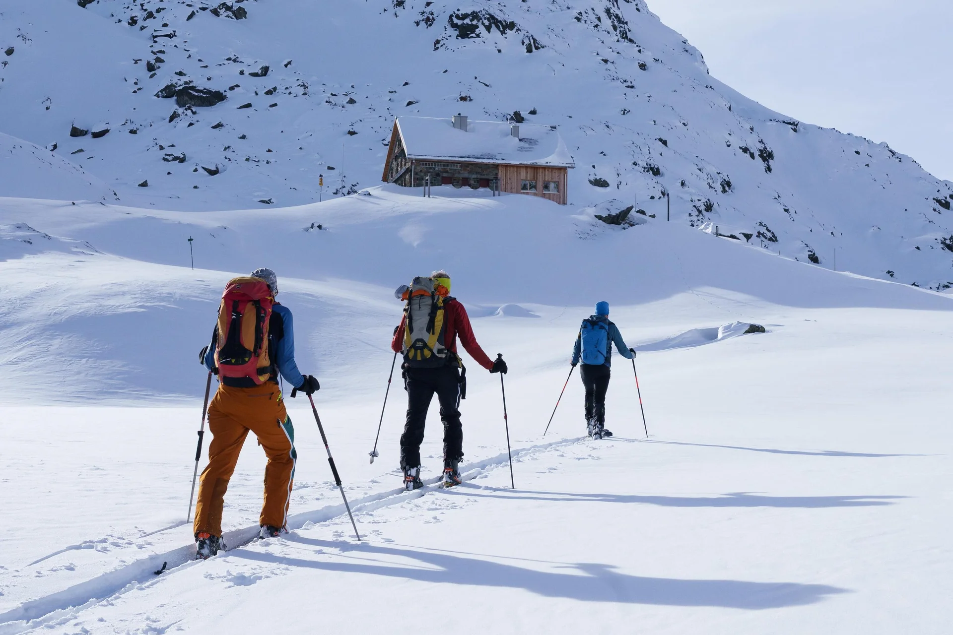 Drei Skitourengeher mit Rucksäcken und Skistöcken wandern in einer verschneiten Berglandschaft auf eine Berghütte zu. | © DAV-FN / B. Rayk