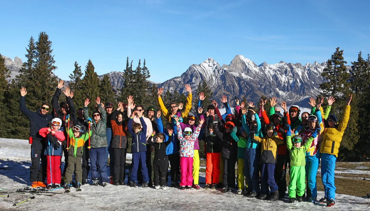 Gruppe von Kindern und Erwachsenen in bunter Winterkleidung steht auf verschneitem Feld vor Bergpanorama und hebt die Arme | © DAV-FN / C. Erbelding