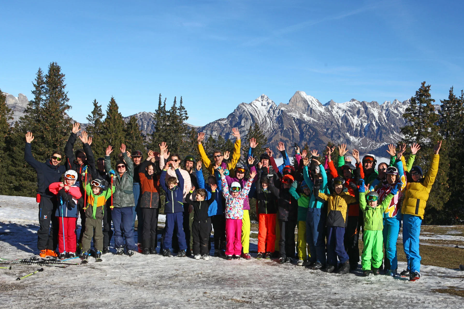 Gruppe von Kindern und Erwachsenen in bunter Winterkleidung steht auf verschneitem Feld vor Bergpanorama und hebt die Arme | © DAV-FN / C. Erbelding