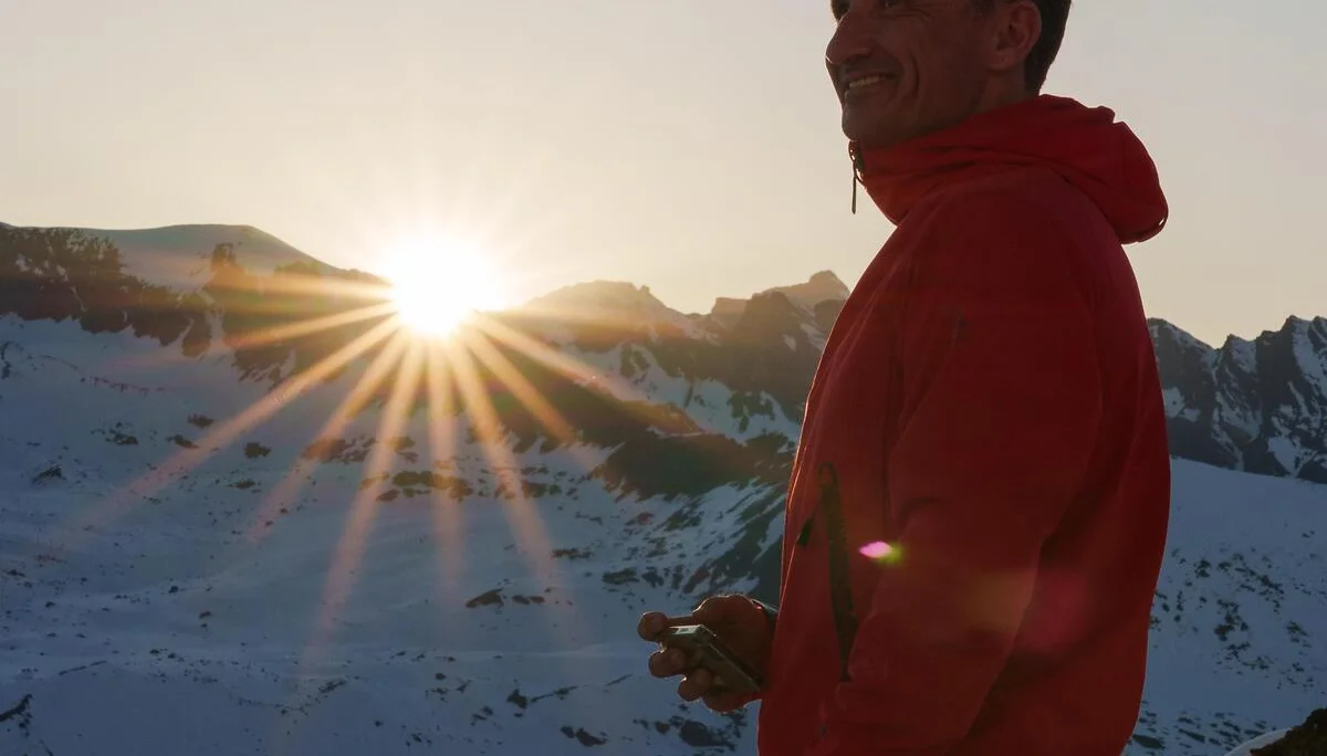 Mann in roter Jacke steht bei Sonnenuntergang vor verschneiten Bergen und hält ein GPS-Gerät in der Hand. | © DAV-FN / R. Gersten u. L. Chr. Fischer