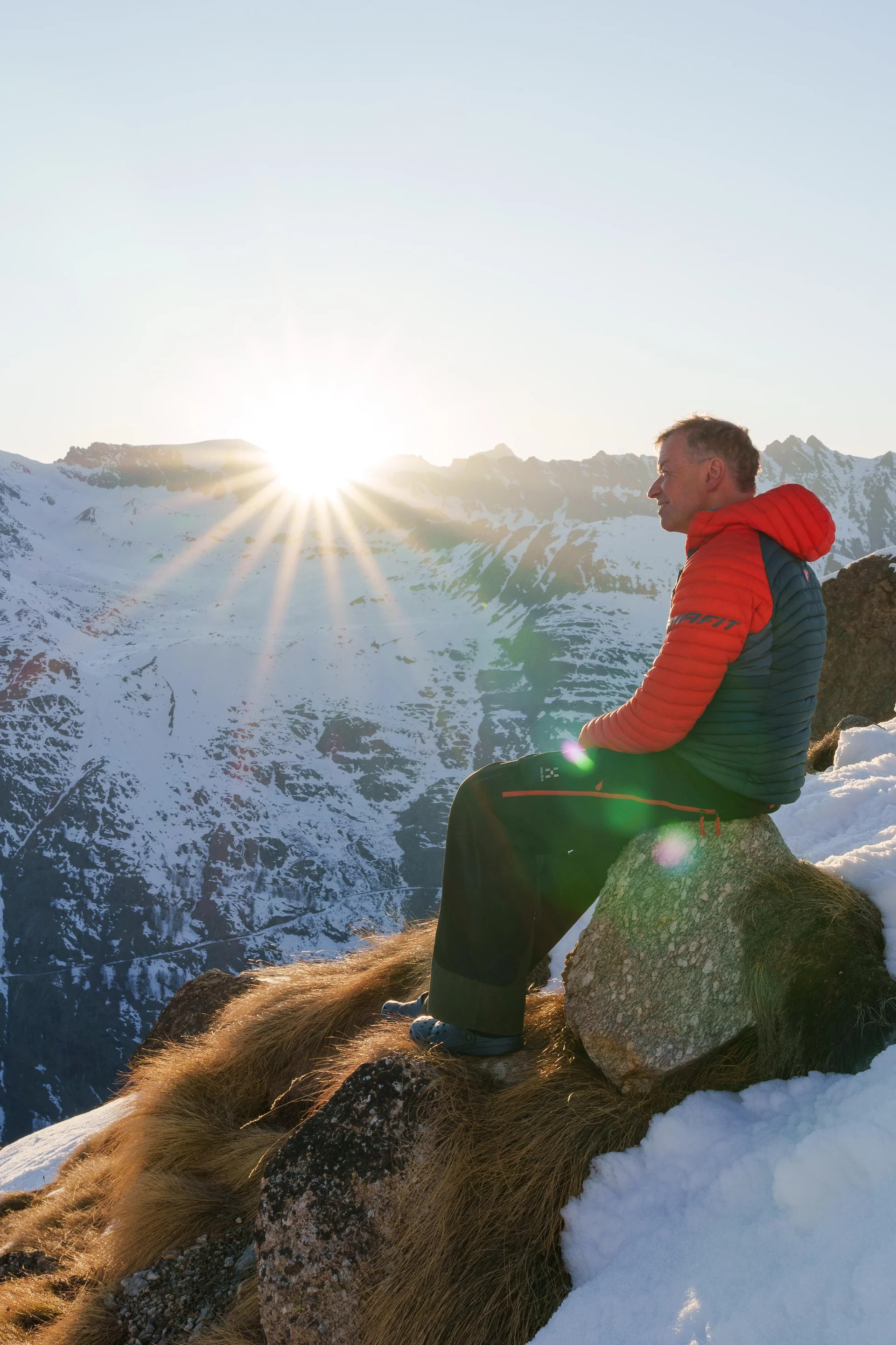 Mann in roter Jacke sitzt auf einem Felsen vor verschneiter Bergkulisse, die Sonne geht hinter den Gipfeln unter. | © DAV-FN / R. Gersten u. L. Chr. Fischer