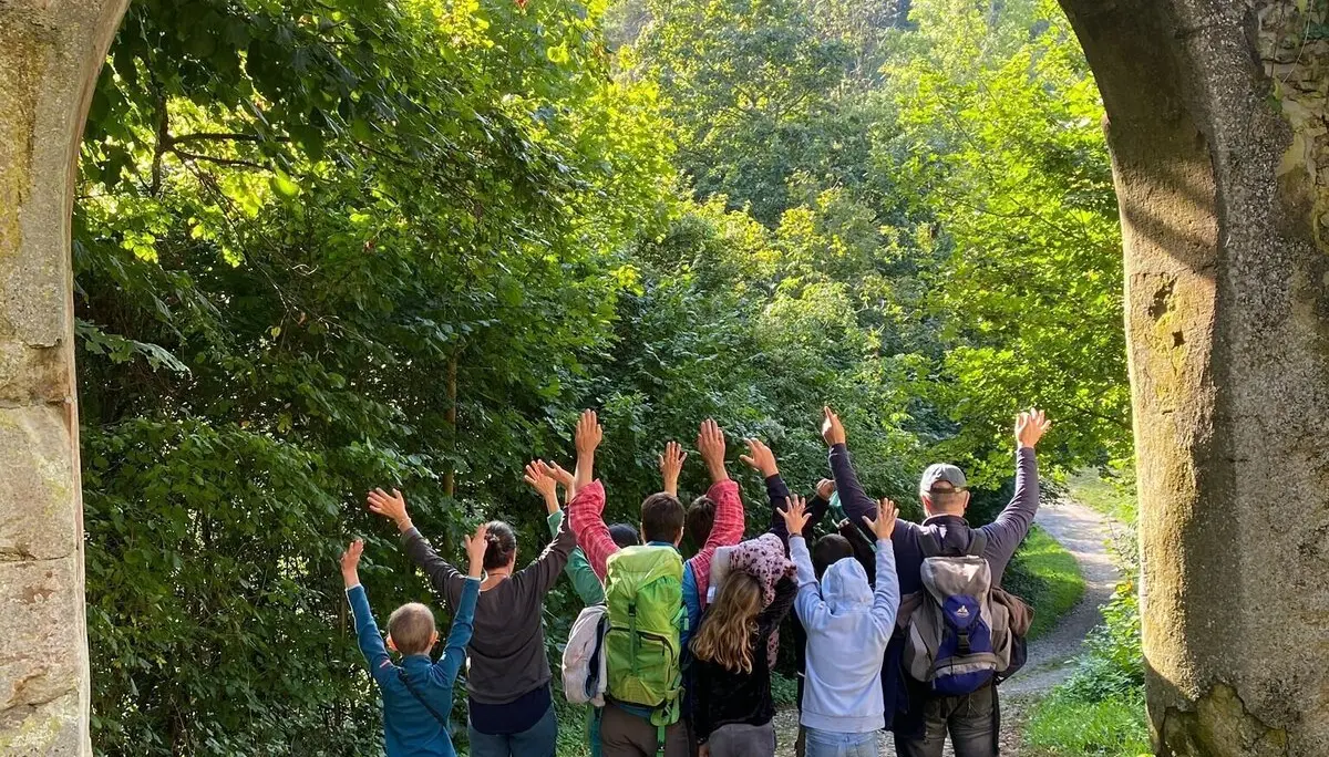 Gruppe von Menschen steht unter einem steinernen Bogen und hebt die Arme in die Luft, umgeben von grünem Laub und einem gepflasterten Weg | © DAV-FN / M. Gross