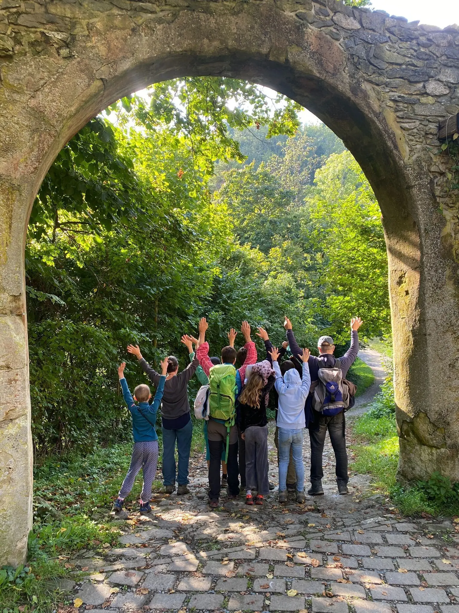 Gruppe von Menschen steht unter einem steinernen Bogen und hebt die Arme in die Luft, umgeben von grünem Laub und einem gepflasterten Weg | © DAV-FN / M. Gross