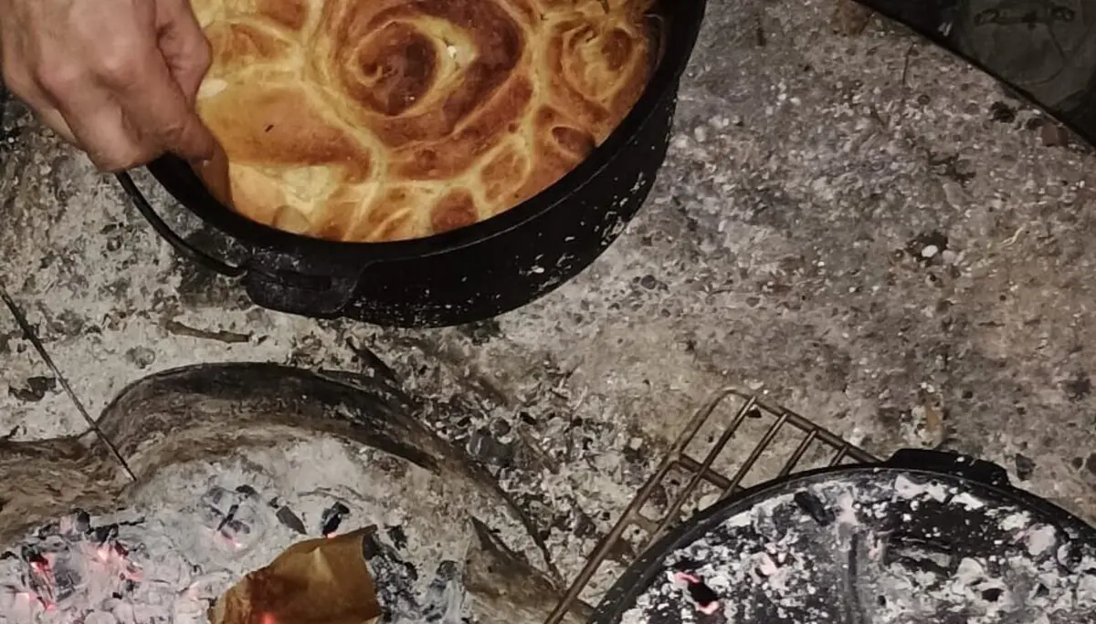 Mehrere Hände nehmen ein rundes, goldbraun gebackenes Brot aus einem schwarzen Topf über einer Feuerstelle mit glühender Asche | © DAV-FN / M. Gross